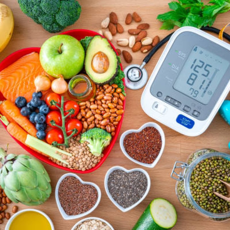 Heart care concept. Overhead view of a group of food that contains healthy nutrients for heart care, a blood pressure monitor, dummbells, tape measure and sthetoscope shot on wooden table. The composition includes extra virgin olive oil, legumes, avocado, berries, nuts and seeds, salmon, broccoli, carrot, soy beans, brown lentils, eggplent, spinach, oats, artichoke, apple, tomato, garlic among others. High resolution 42Mp studio digital capture taken with SONY A7rII and Zeiss Batis 40mm F2.0 CF lens